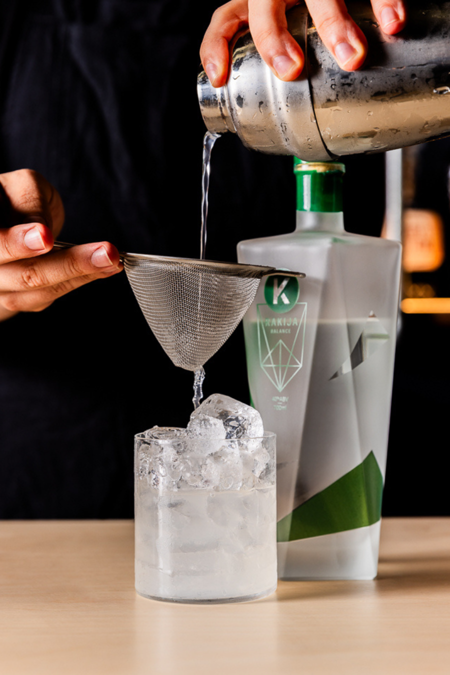 A mixologist pouring a cocktail shaker with a rakija cocktail through a strainer and into a glass of ice.