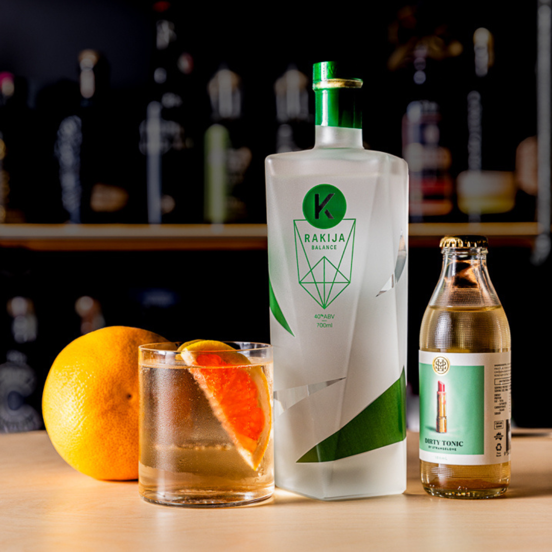 A Rakija & Tonic mixed drink, garnished with orange, next to a bottle of Kabina Balance Rakija and Strangelove Dirty Tonic 
