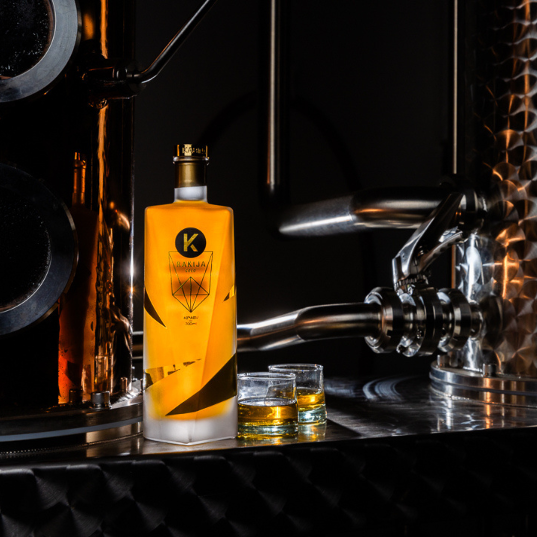A bottle of Kabina Gold Rakija 40% ABV, with two shot glasses of Kabina gold positioned beside the bottle. A copper still is behind the bottle.