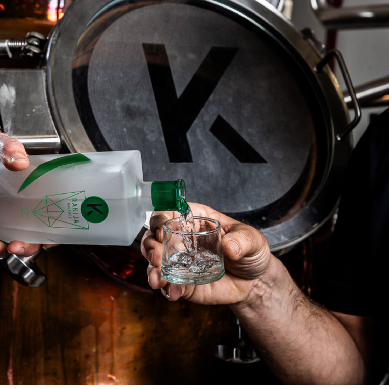 A copper kettle embossed with the letter K for Kabina Distilling, in which rakija is distilled in Melbourne, Australia. A person is pouring a shot glass of Kabina Balance rakija in front of the copper kettle.